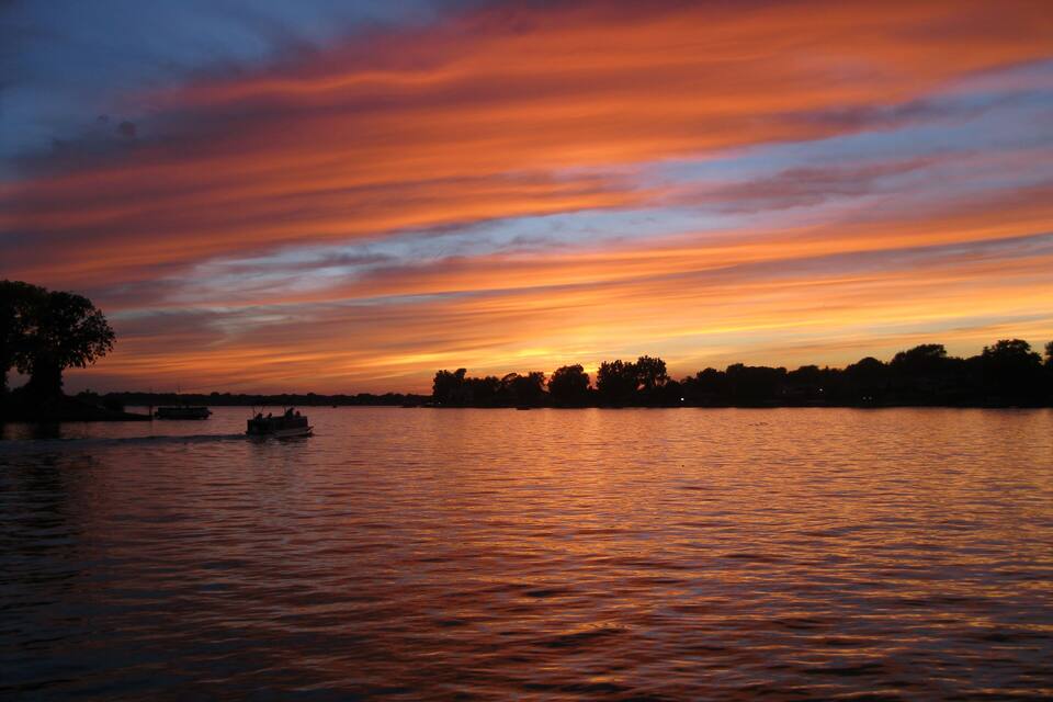 Een zonsondergang boven een kalm meer, met bootjes en bomen in silhouet tegen de lucht.