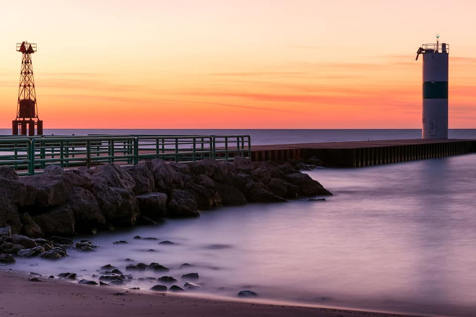 A coastal sunset with a pier, a lighthouse, and rocky shoreline.