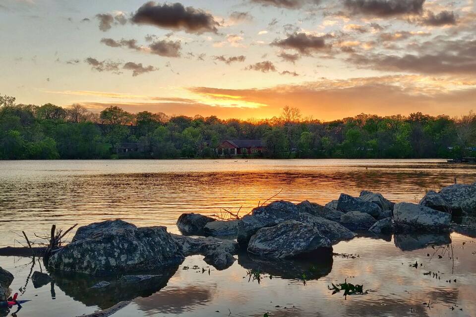 Un lago tranquilo al atardecer, con rocas en primer plano y árboles al fondo.