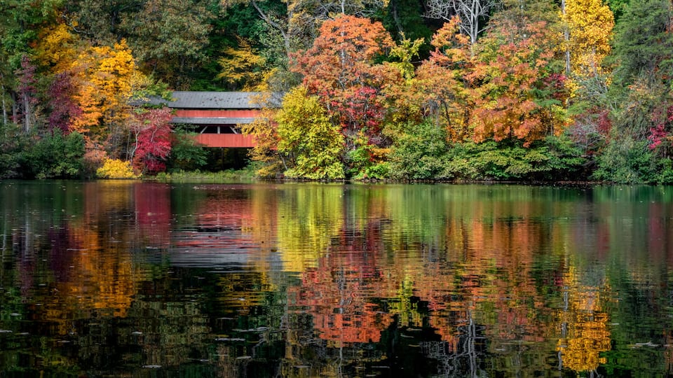 With a beautiful reflection on Lake Loretta in Alley Park, Lancaster, Ohio, the red George Hutchins Covered Bridge, surrounded by colorful autumn leaves, was constructed in 1865 at another location.