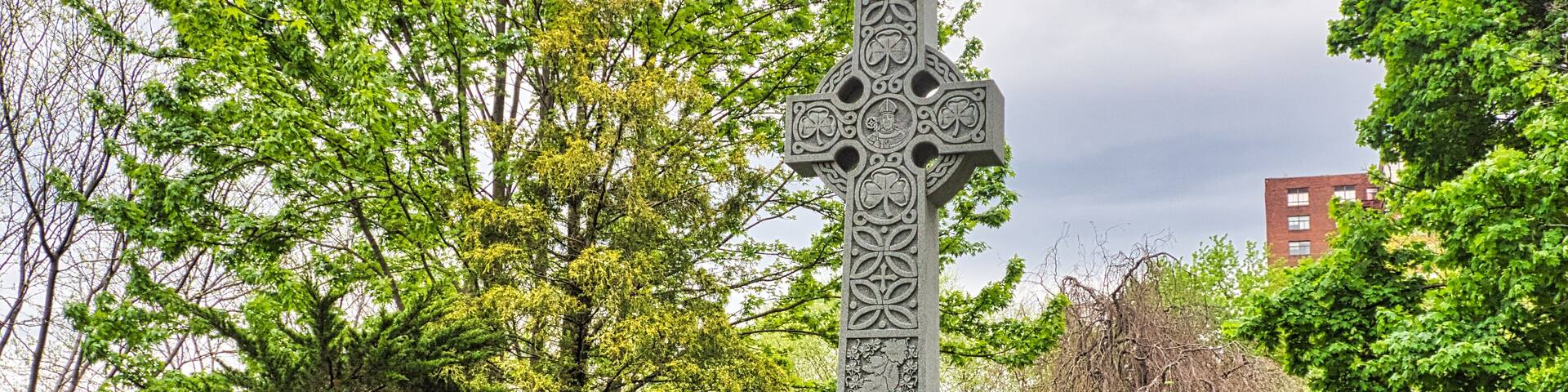 A stone cross with intricate carvings, surrounded by lush greenery and trees.