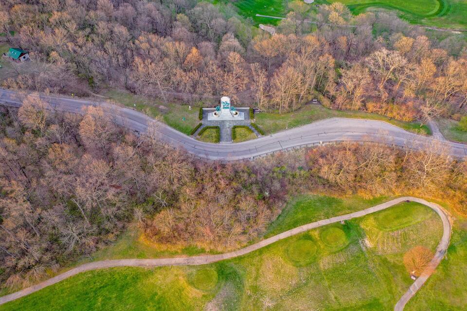 An aerial view of a park with a central monument, winding paths, and surrounding trees.