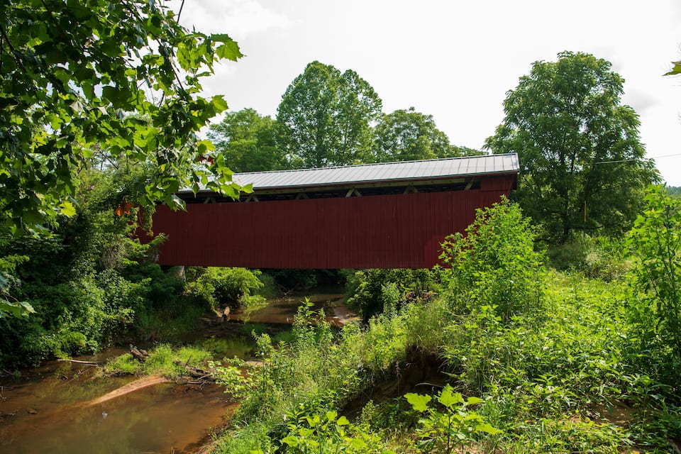 Un puente cubierto de color rojo cruzando un arroyo pequeño, rodeado de vegetación exuberante.