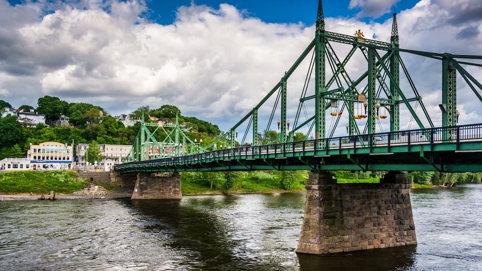 A green steel bridge over a river with buildings and greenery in the background.
