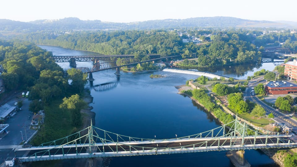 A suspension bridge over a river with surrounding greenery and a distant town.