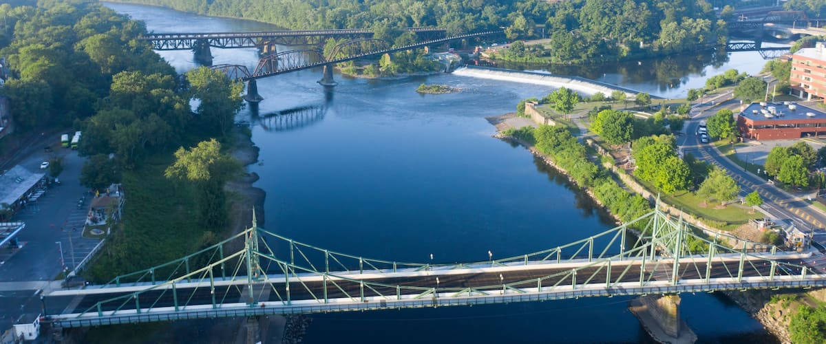 A suspension bridge over a river with surrounding greenery and a distant town.