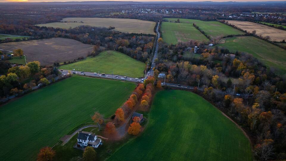 An aerial view of a rural landscape with fields, a winding road, and a small house surrounded by trees.