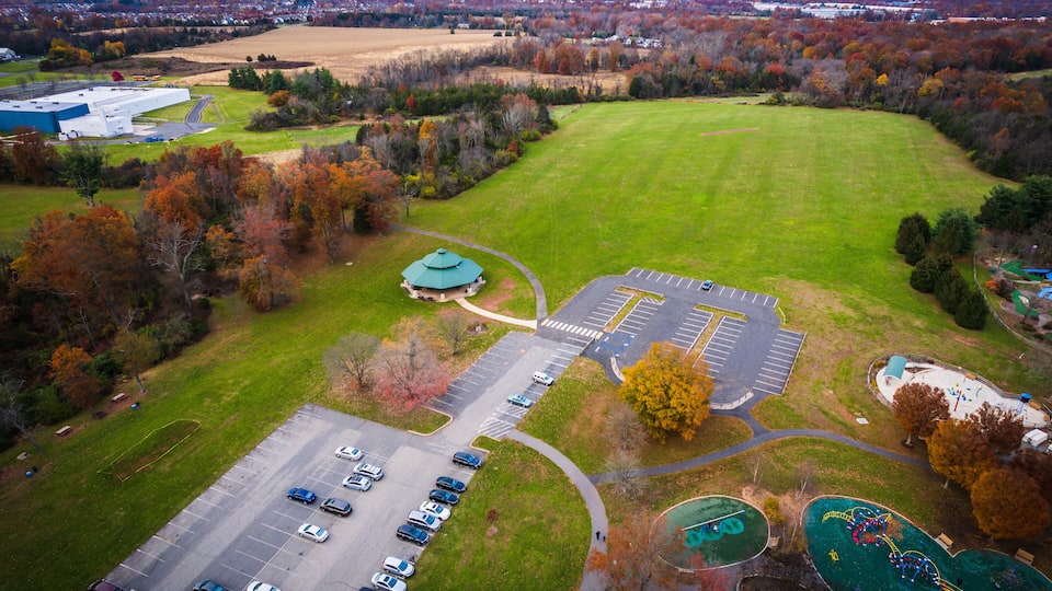 An aerial view of a park with a parking lot, playgrounds, and surrounding autumn-colored trees.