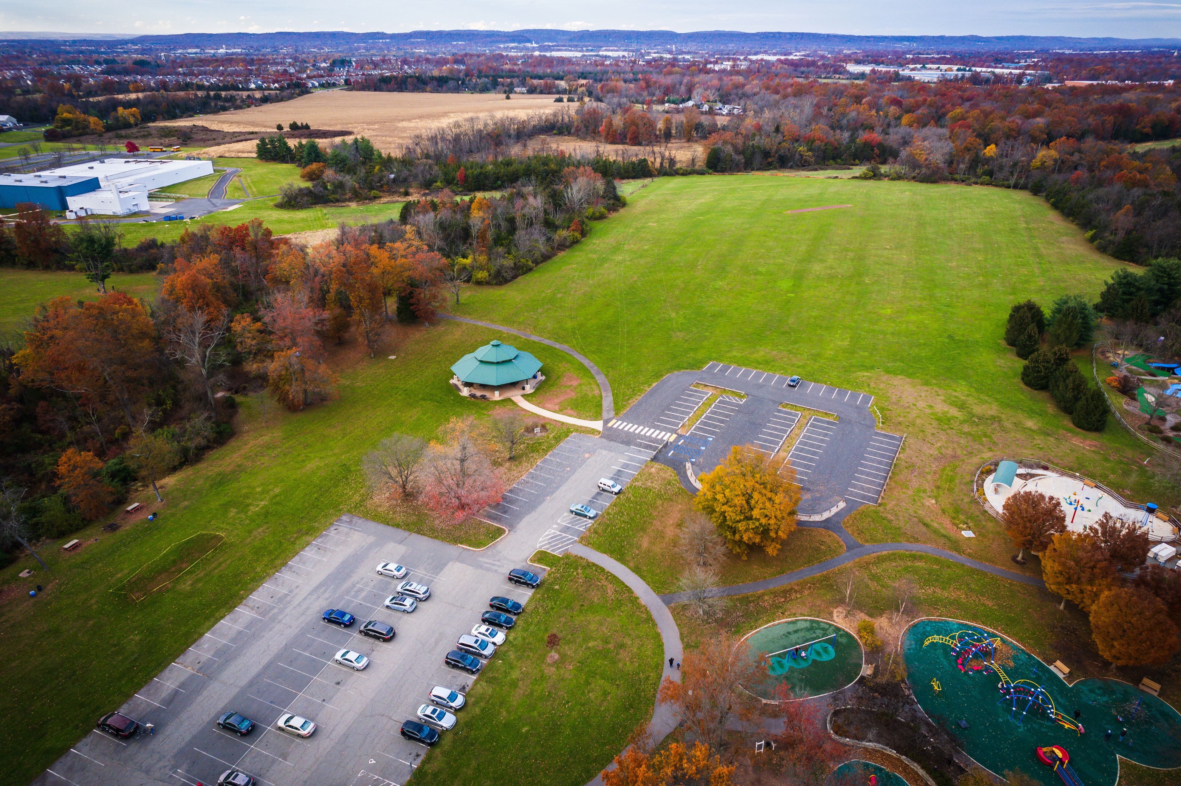An aerial view of a park with a parking lot, playgrounds, and surrounding autumn-colored trees.