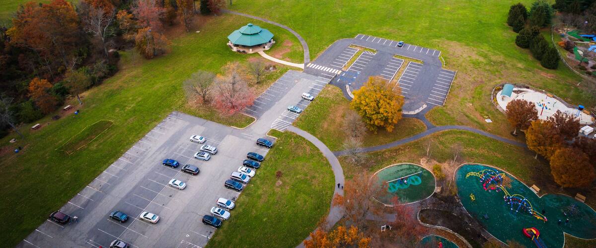 An aerial view of a park with a parking lot, playgrounds, and surrounding autumn-colored trees.