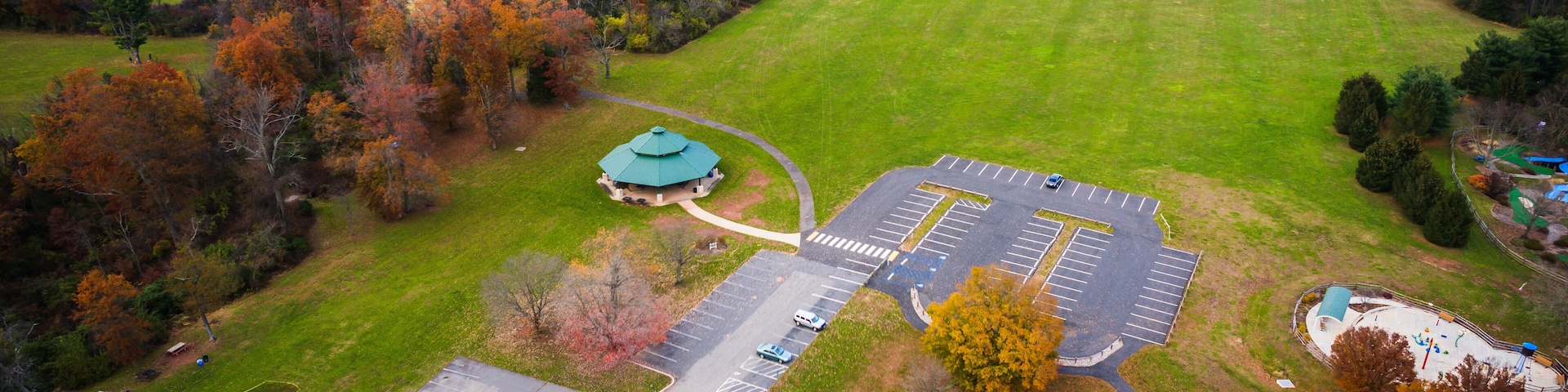 An aerial view of a park with a parking lot, playgrounds, and surrounding autumn-colored trees.