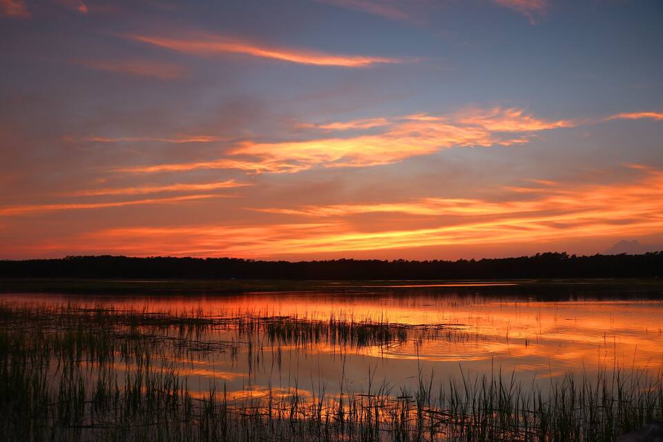 A sunset over a calm lake with reflections of the sky, surrounded by tall grasses.