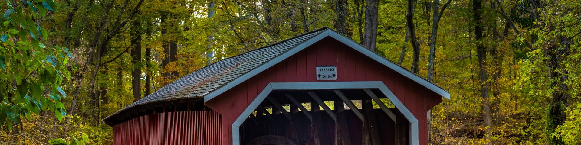 Un pont couvert de couleur rouge, avec un mur de pierre d’un côté et une clôture en bois de l’autre, le tout entouré d’un feuillage automnal.
