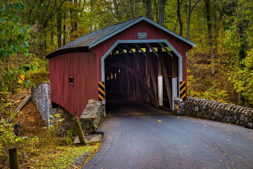 A red covered bridge with a stone wall on one side and a wooden fence on the other, surrounded by autumn foliage.