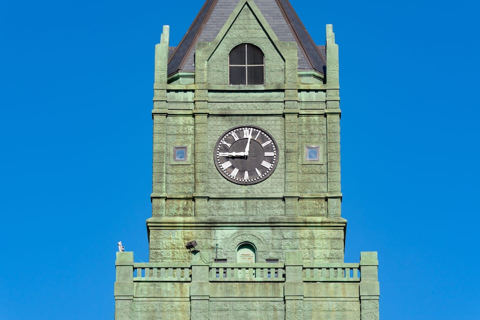 A clock tower with a greenish stone facade, a pointed roof, and a clear blue sky in the background.