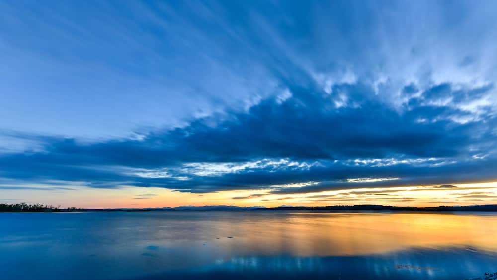 Un lac paisible dont la surface reflète le ciel, bordé d’une rive rocheuse et entouré de montagnes lointaines.