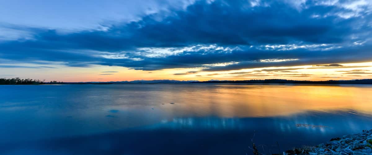 A calm lake with a reflection of the sky, surrounded by a rocky shoreline and distant mountains.
