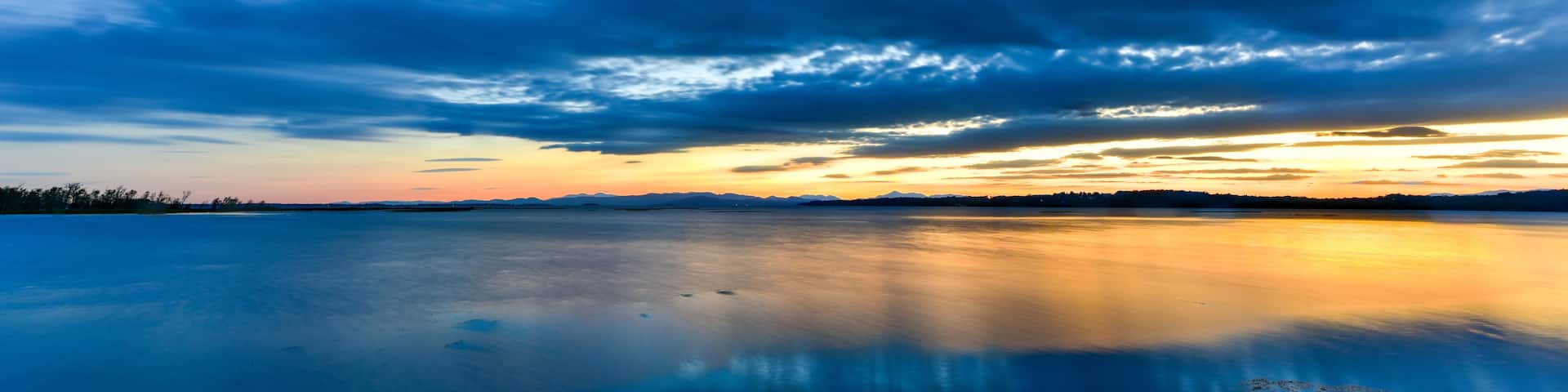 A calm lake with a reflection of the sky, surrounded by a rocky shoreline and distant mountains.