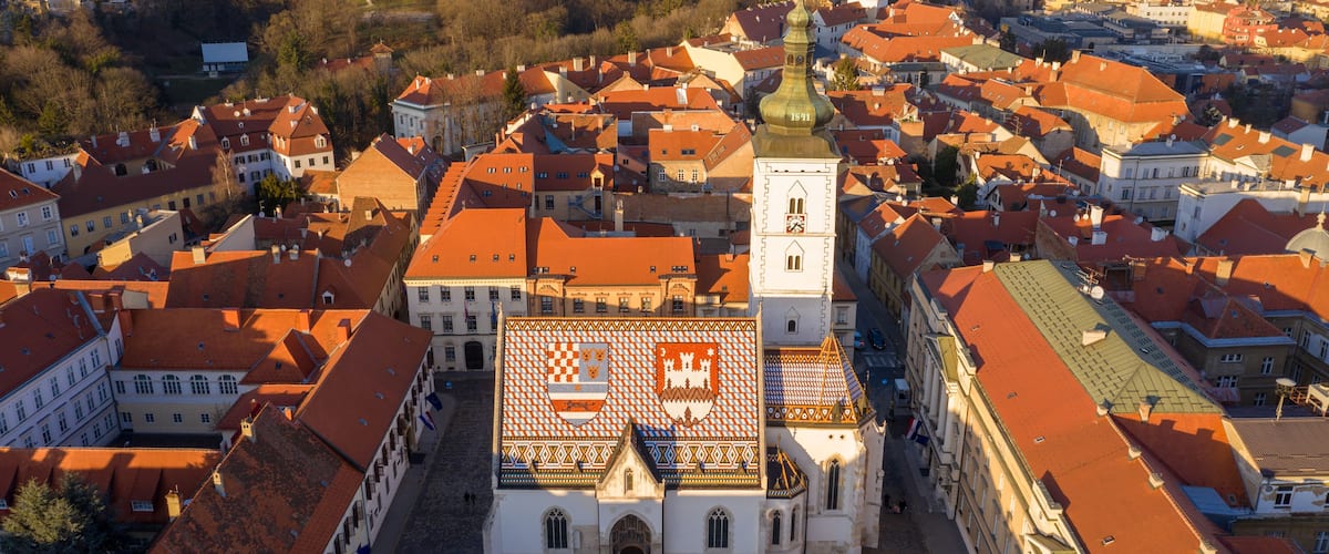 A historic town with red-roofed buildings, a prominent church with a clock tower, and a large square with a patterned roof.