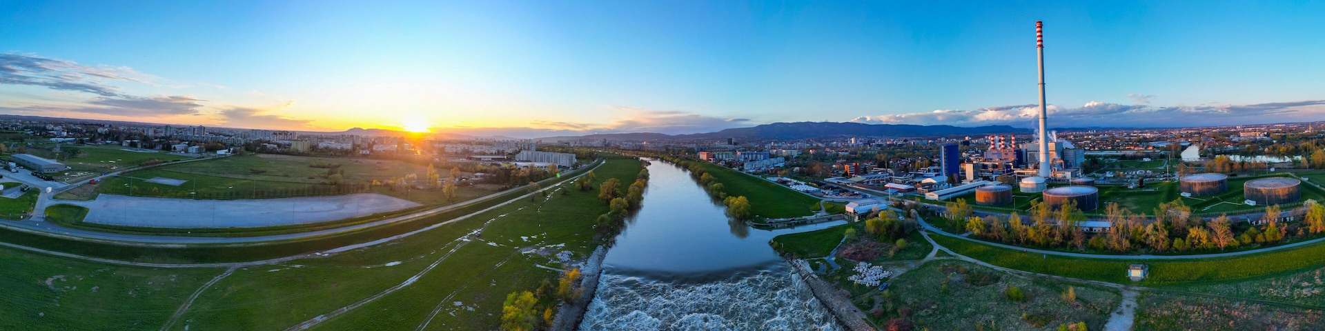 Une rivière traverse une ville où se trouvent des bâtiments industriels et une haute cheminée d’usine.