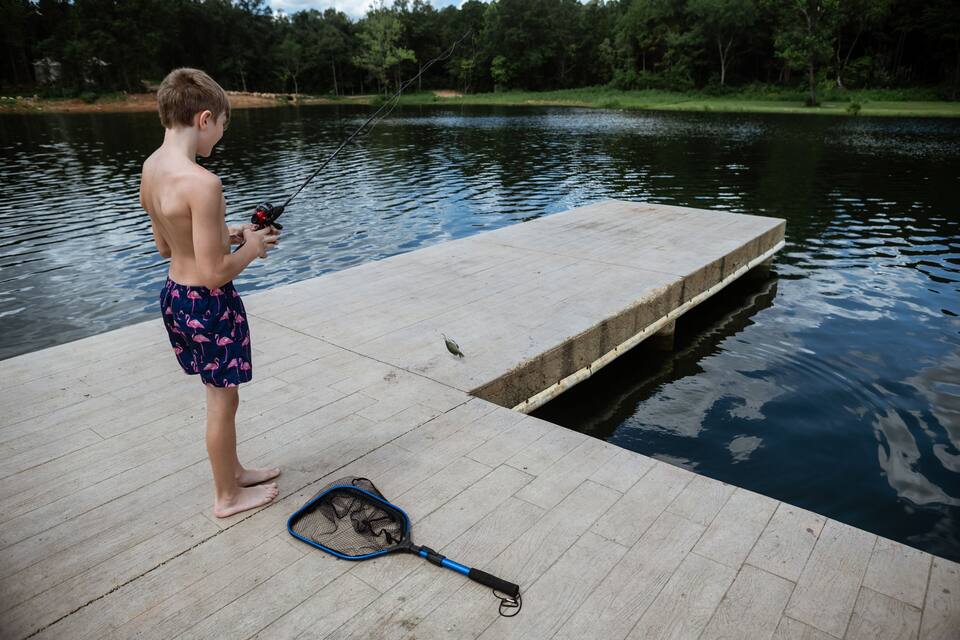 Un garçon pêche sur un quai en béton, au bord d’un lac paisible entouré d’arbres.