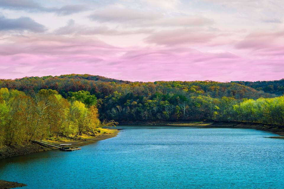 A river with a wooden dock, surrounded by autumn-colored trees and hills in the background.