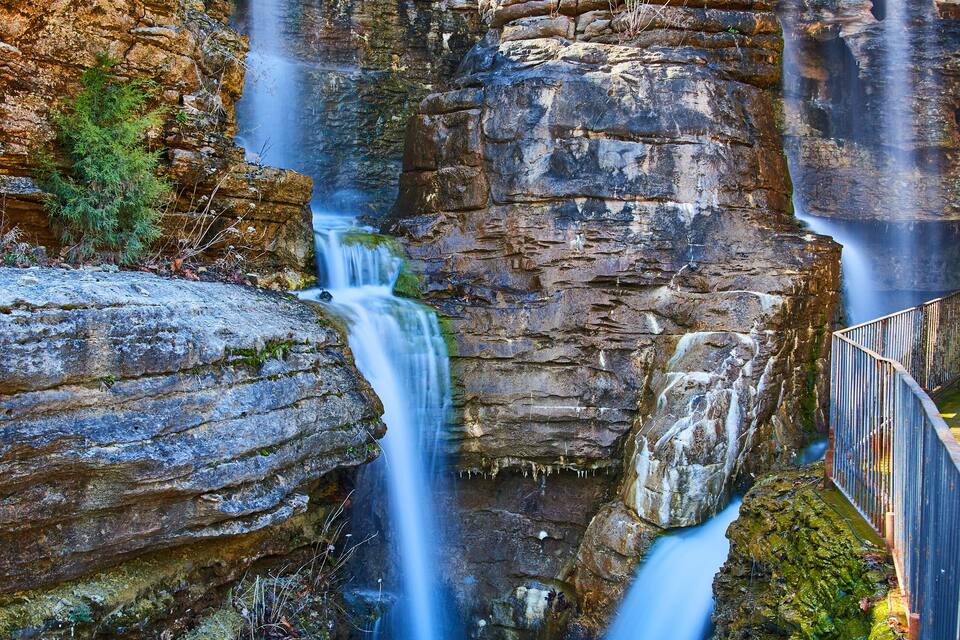 A multi-tiered waterfall cascading over rocky cliffs with a wooden walkway nearby.