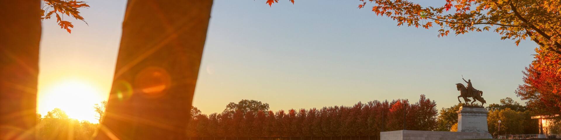 A statue in a park with autumn leaves and a sunset in the background.