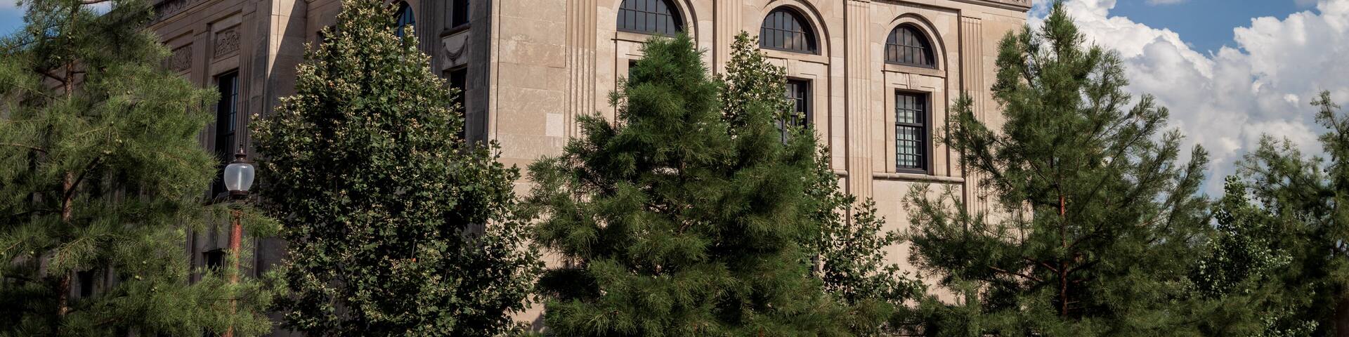 Un bâtiment historique doté d’arcs en plein cintre et surmonté d’une tour horloge, entouré d’arbres.