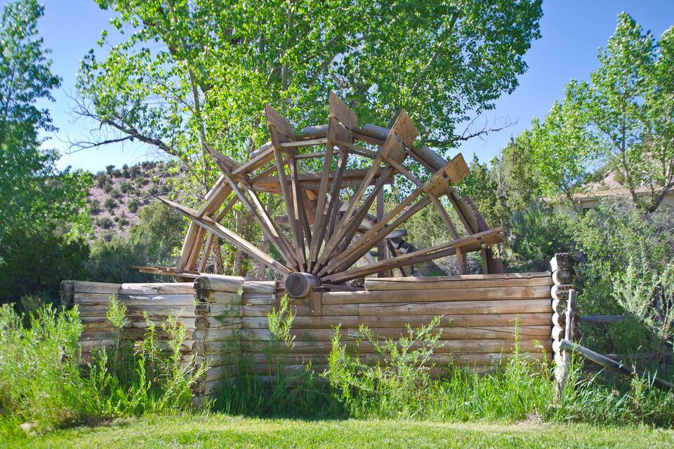 A wooden water wheel in a natural setting with surrounding greenery and hills.