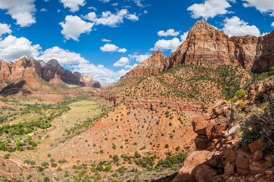 A canyon with red rock formations, a valley with sparse vegetation, and a clear blue sky with clouds.