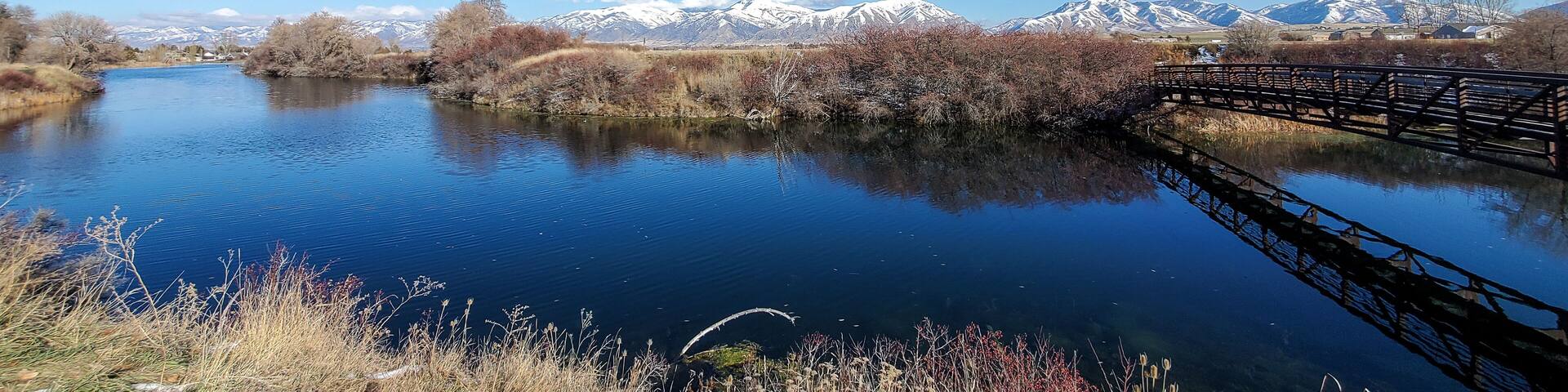 A calm river with a wooden bridge, snow-capped mountains in the background, and autumn-colored trees.