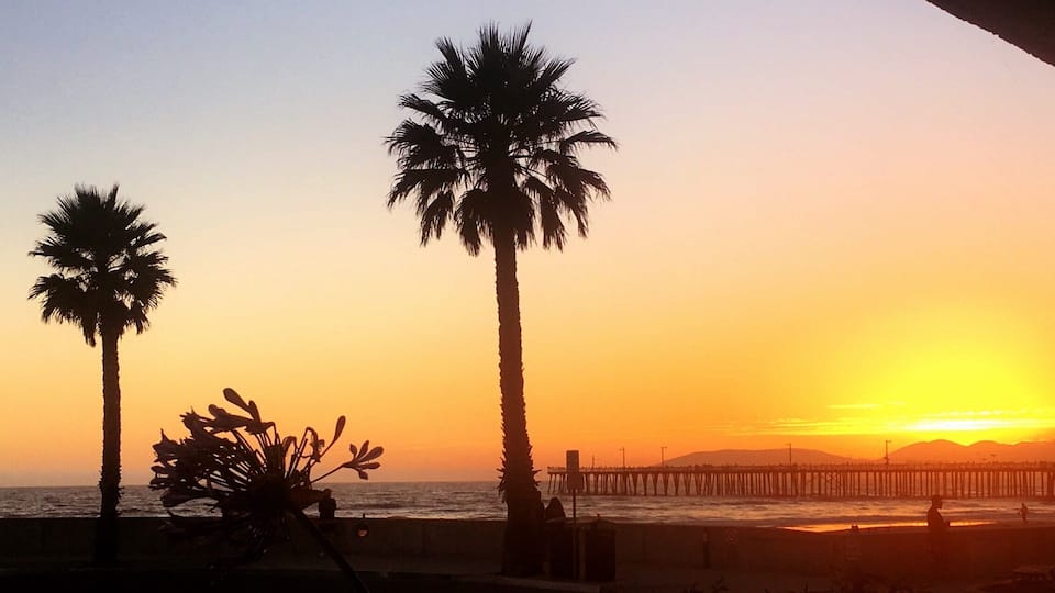 A sunset over a beach with palm trees and a pier.