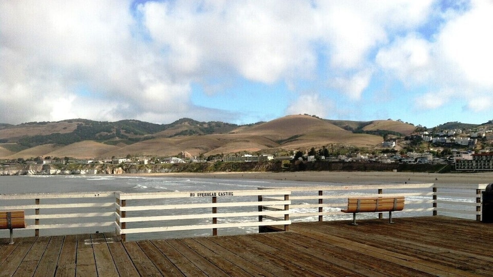 A wooden pier with benches overlooking a beach with a backdrop of hills and a town.