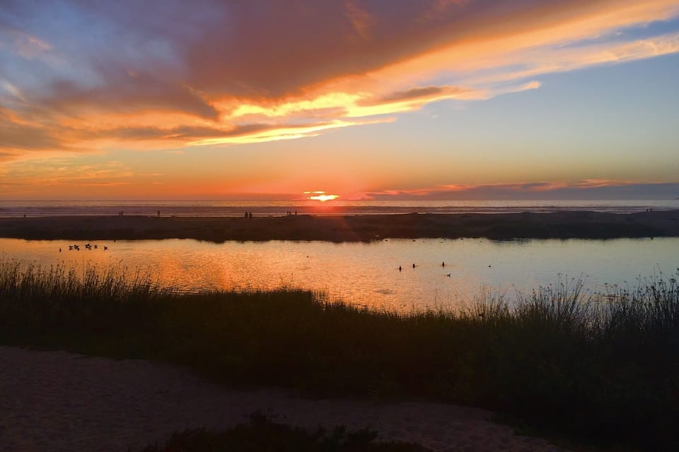 Un atardecer sobre un cuerpo de agua tranquilo, con siluetas de personas y aves.