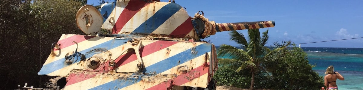 A rusted military tank with a colorful striped pattern on a sandy beach near the ocean.