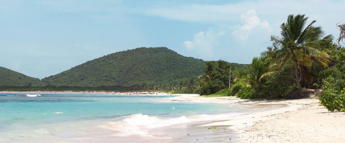 A beach with clear turquoise water, white sand, and lush green hills in the background.