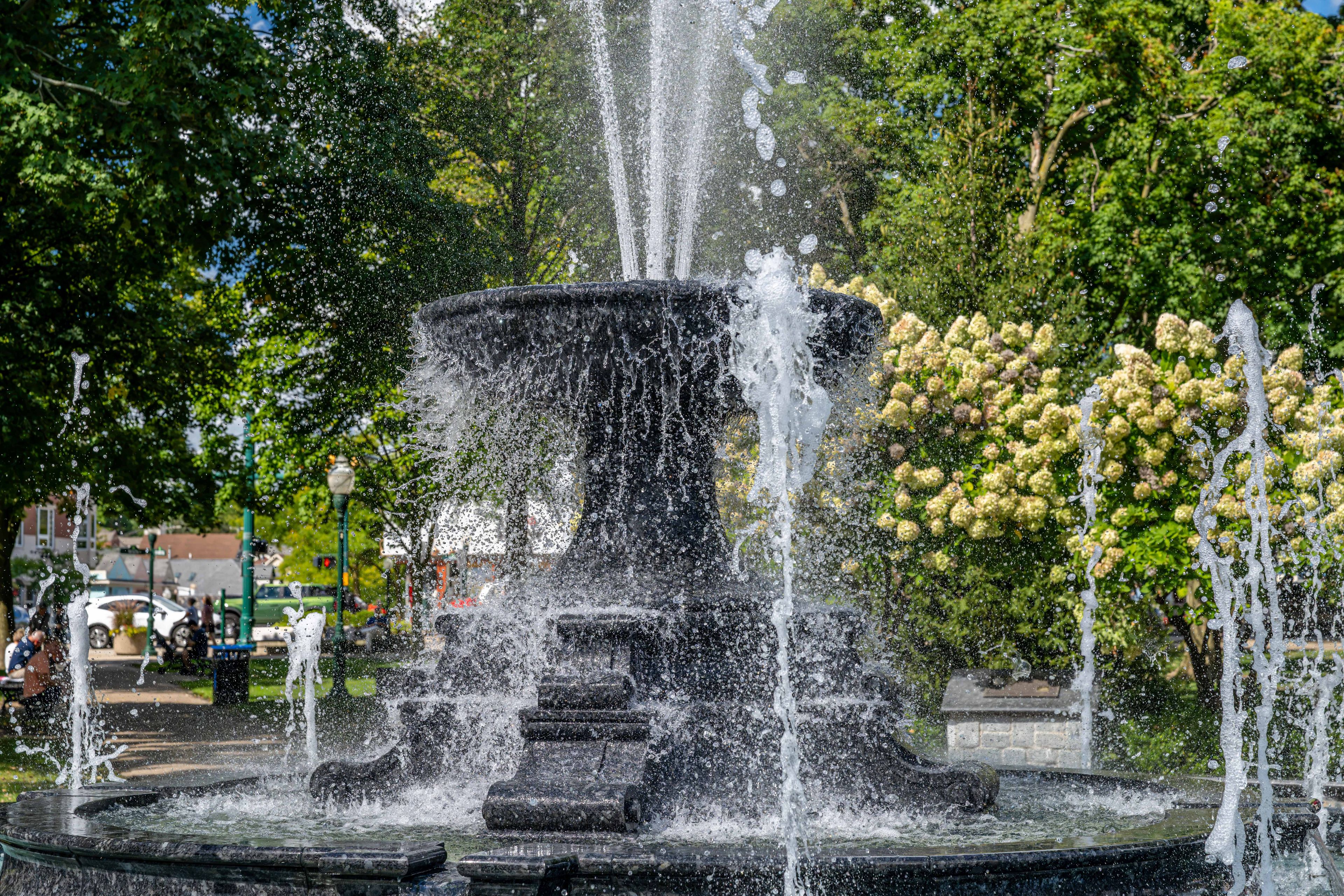 Een fontein met verschillende waterstralen in een park met bomen en een gebouw op de achtergrond.