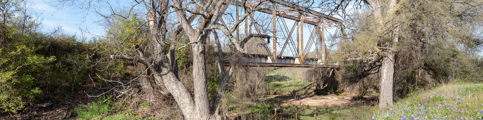 A rusted bridge over a stream, surrounded by trees and wildflowers.