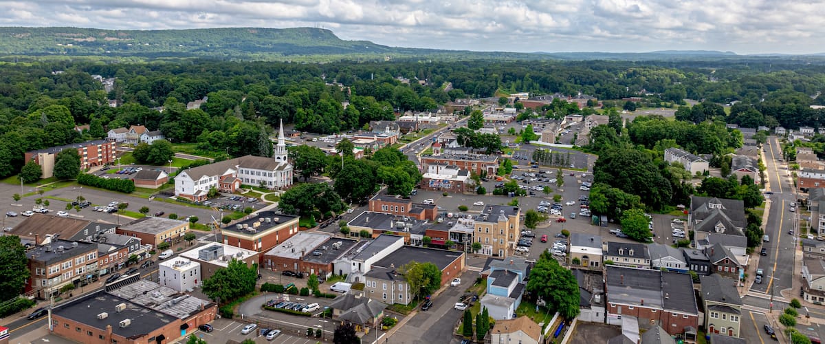 A town with a prominent church steeple, a mix of residential and commercial buildings, and a backdrop of green hills.
