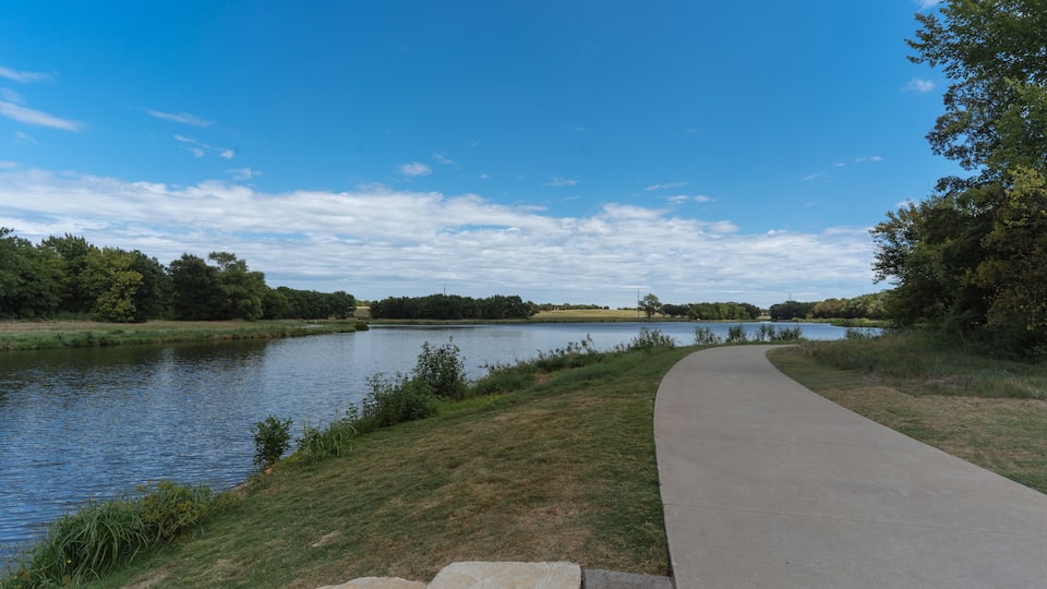A paved walkway beside a calm river, surrounded by grassy banks and trees under a clear sky.