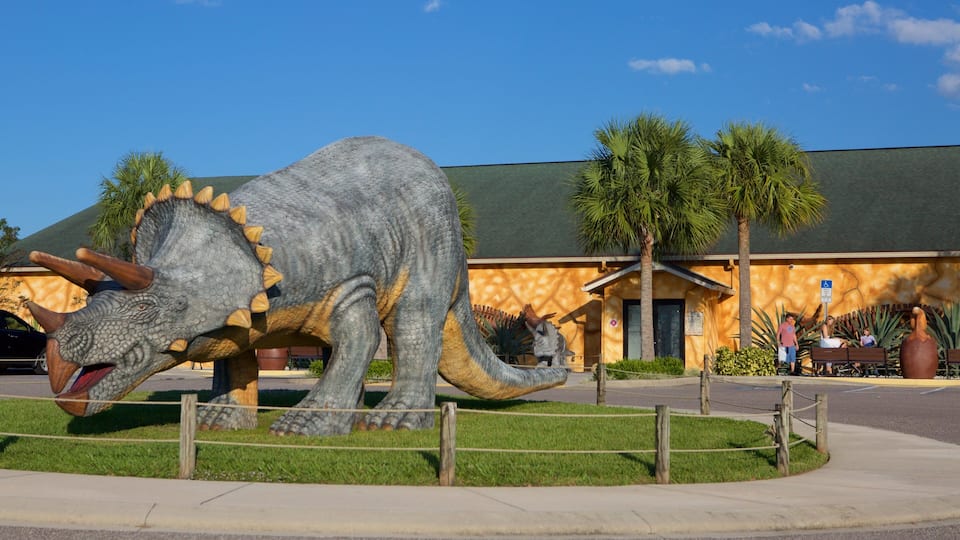 A large dinosaur statue in a fenced area with palm trees and a building in the background.