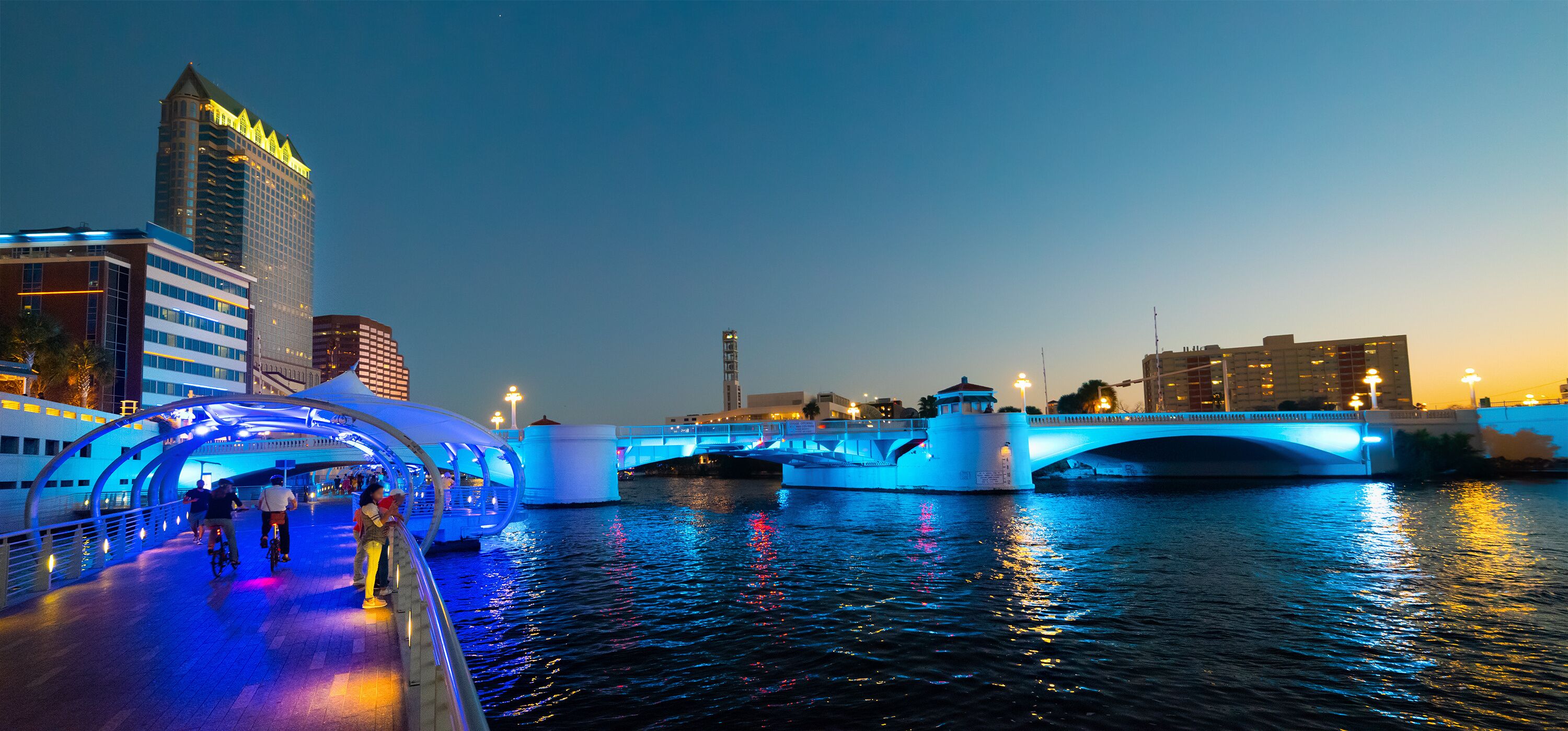 A cityscape at dusk with illuminated bridges over a river, modern buildings, and a prominent skyscraper.