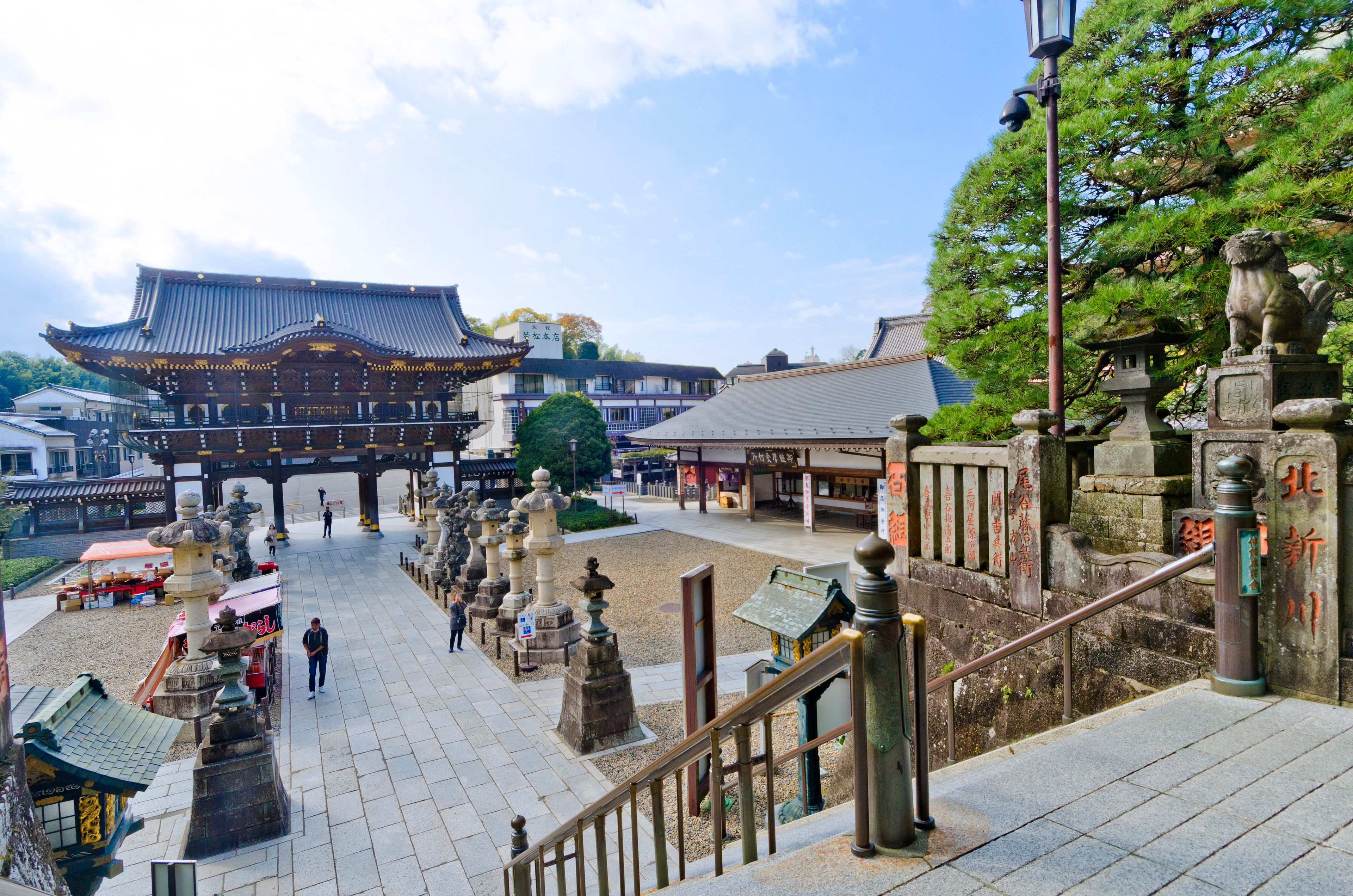 Une porte traditionnelle japonaise, dotée d’une architecture richement décorée, une lanterne en pierre et un escalier menant à celle-ci.