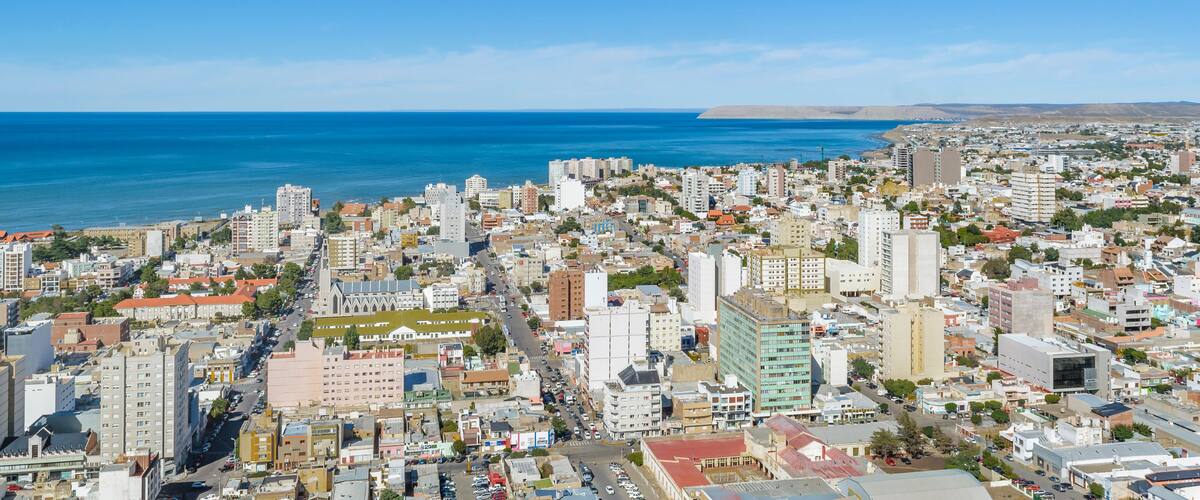 A coastal cityscape with dense urban buildings, a clear blue sky, and the ocean in the background.