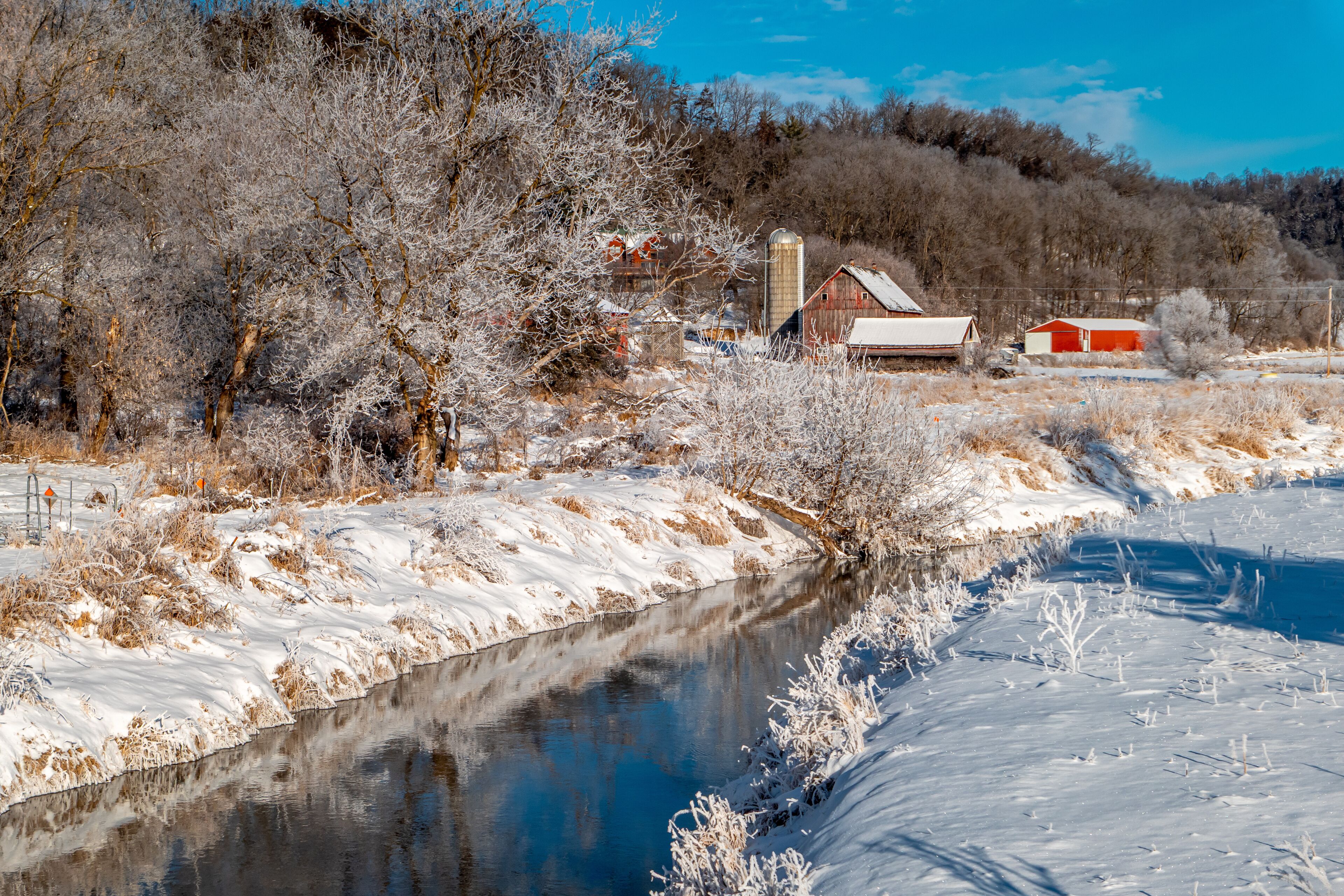 A snow-covered riverbank with a red barn and a silo in the background.