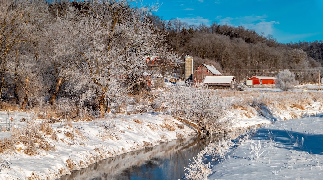 A snow-covered riverbank with a red barn and a silo in the background.