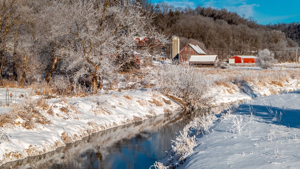 A snow-covered riverbank with a red barn and a silo in the background.