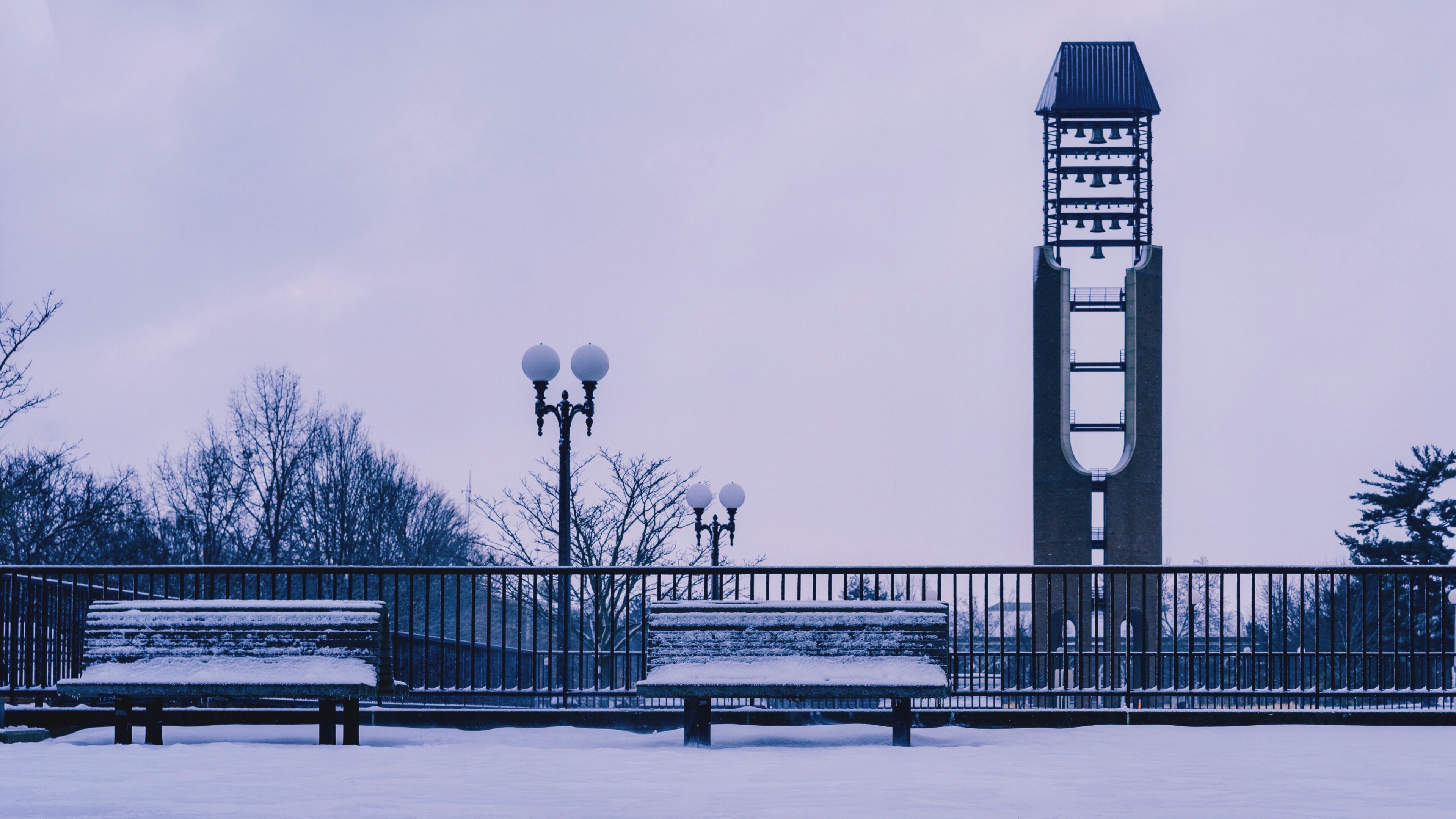 A snow-covered bench, a metal fence, and a tower with a bell structure.