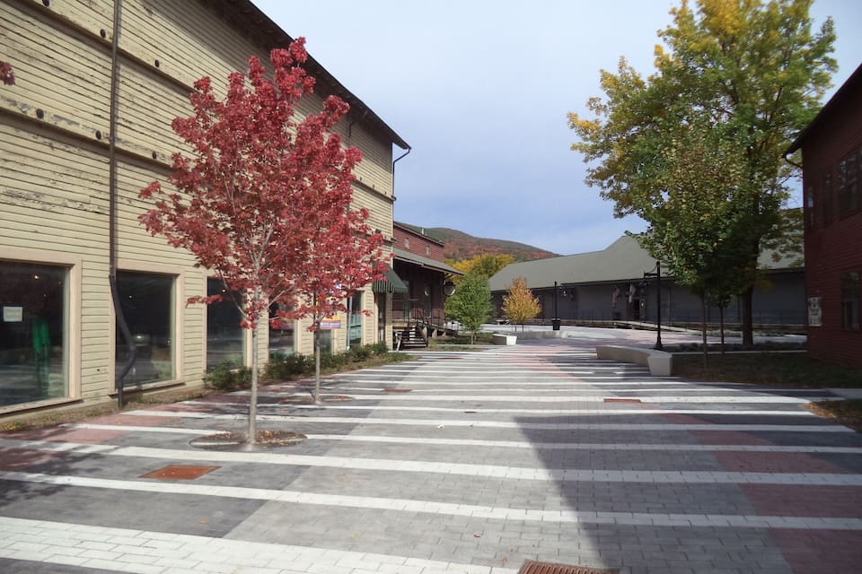 A pedestrian plaza with a crosswalk, trees with autumn foliage, and buildings with commercial signage.